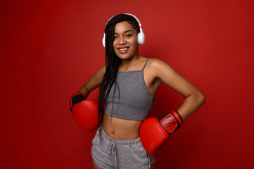 Athlete African woman boxer in wireless headphones and hands in boxing gloves on waist, smiles...