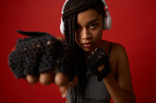 Close-up of concentrated young African athletic woman boxer in headphones and red boxing gloves making a straight kick, isolated on color background with copy space. Contact martial art concept - Powered by Adobe