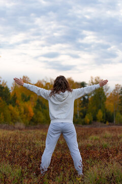 A woman in a white sweater with a hood stands in a field against the background of a forest and a blue sky with her arms outstretched wide on a warm autumn day. Selective focus. Close-up. Portrait