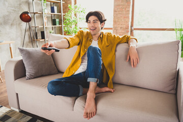Photo of positive handsome guy hold remote control watch tv series sit couch wear yellow shirt home...