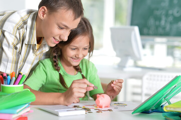 Cute boy and girl pitting coins into piggy bank while sitting at table