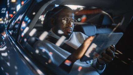 Stylish Black Man in Glasses is Commuting Home in a Backseat of a Taxi at Night. Male Using Tablet Computer and Looking Out of Window while in a Car in Urban City Street with Working Neon Signs.