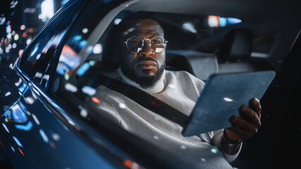 Stylish Black Man in Glasses is Commuting Home in a Backseat of a Taxi at Night. Male Using Tablet...