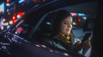 Stylish Female is Commuting Home in a Backseat of a Taxi at Night. Beautiful Woman Passenger Using Smartphone while in a Car in Urban City Street with Working Neon Signs.
