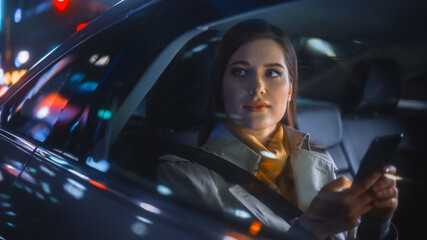 Stylish Female is Commuting Home in a Backseat of a Taxi at Night. Beautiful Woman Passenger Using Smartphone and Looking Out of Window while in a Car in Urban City Street with Working Neon Signs.