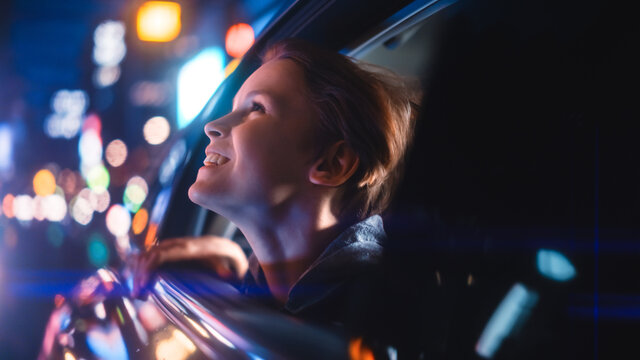 Excited Young Boy Is Sitting On Backseat Of A Car, Commuting Home At Night. Looking Out Of The Window With Amazement Of How Beautiful Is The City Street With Working Neon Signs.