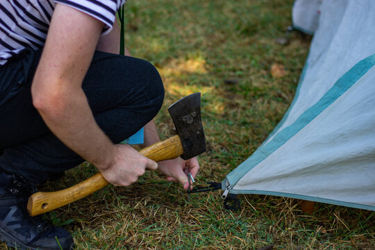 Male camper pitch a tent in the forest. Man helps set up. Man driving tent stakes with axe. Person stretching tent stakes. Getting ready to camping. Person setting up tent. Hammering down a nail.