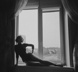 A beautiful girl in a bodysuit sits on the window, black and white photo.