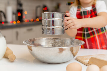 little happy smiling blonde girl in a chef costume kneads dough for Christmas baking in the kitchen. kid is preparing a festive dinner. child sifts the flour through a sieve. new year's winter concept
