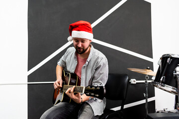 Young man with beard in Santa hat is playing Christmas music on acoustic guitar.