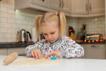 cute little blonde girl with colorful shapes cuts out cookies from the dough. little cook prepare Christmas pastry in the kitchen. festive breakfast, lunch, dinner. space for text. High quality photo