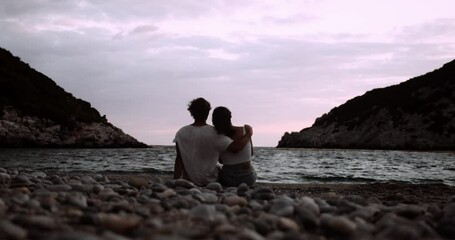 happy caucasian couple sitting on beach arms around each other at sunset
