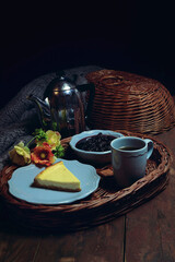 Afternoon tea. on a wicker tray is a plate of cheesecake and jam. In the background, a teapot with boiling water and a mug of tea.