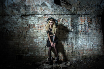 A woman in the image of a witch in a bright costume with feathers on Halloween night on the background of an old brick wall of the dungeon.