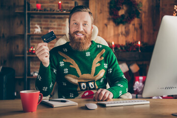 Photo of positive bearded guy sit table demonstrate credit card wear ugly deer sweater decorated...