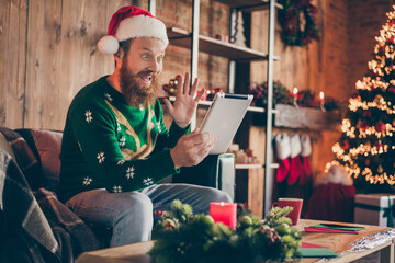 Photo of friendly guy sit armchair hold tablet wave hand video meeting wear santa hat sweater decorated home indoors