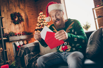 Photo of excited glad impressed guy open envelop take letter wear santa hat pullover decorated home indoors