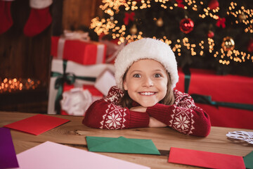 Photo of cute dreamy schoolgirl dressed red sweater headwear writing christmas congratulations indoors room home house