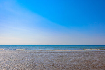 The waves of the ocean break onto the wet sandy beach, overlooked by a beautiful and clear blue sky