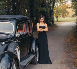 Vintage style photo. Elegant retro woman in black evening long dress posing on road, autumn nature...