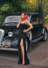 Vintage style photo. Elegant retro woman in black evening long dress posing on road autumn nature background green orange leaves trees. Old car. Red lips shoes headscarf. Beautiful face bright makeup
