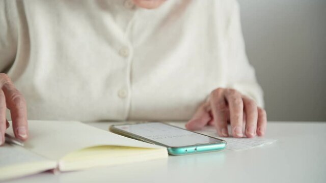 An elderly woman in room puts on glasses, writes in a notebook with a pen and calculates taxes and expenses on a calculator in a smartphone. Concept of budget calculation and pensioners' finances