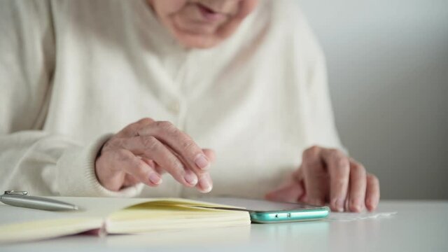 Elderly woman sitting at table, counting on calculator in smartphone, wrinkled hands close-up. Notepad, pen and receipts on table. Concept of budget, tax and pension calculation