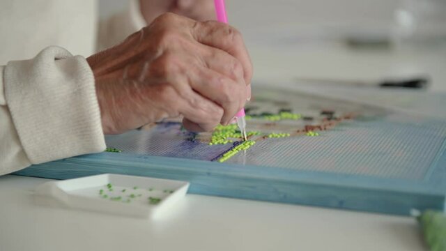 Elderly woman spends free time laying out carefully diamond embroidery using special tool on pre-glued canvas extreme closeup