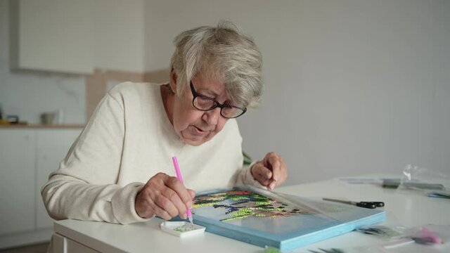 Elderly woman with short gray hair dressed in white jacket takes off eyeglasses and rubs eyes wearily at laying out diamond mosaic
