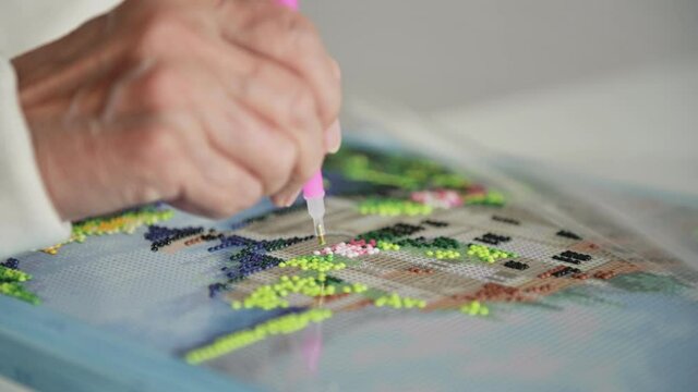 Elderly woman wrinkled hand lays out diamond mosaic embroidery on canvas with special tool spending spare time on retirement macro