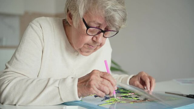 Elderly grey-haired woman with glasses in white woolen jacket lays out diamond mosaic embroidery during spare hobby time at table