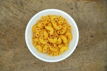 Raw elbow Macaroni (Gomiti Pasta) in white bowl on wooden table with copy space, Table top view