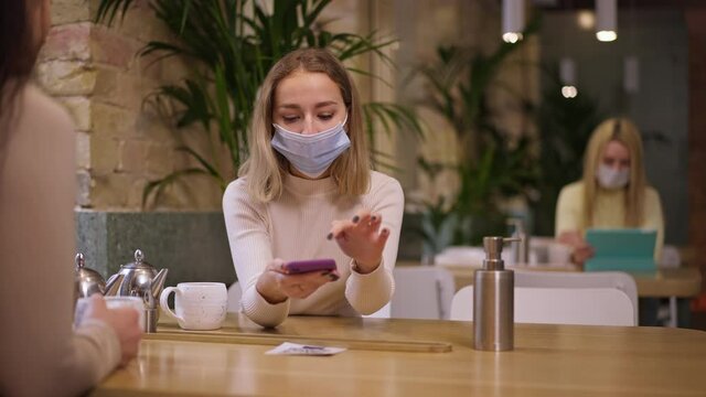 Focused young woman in coronavirus face mask scanning QR code with online menu in cafe sitting at table with friend. Portrait of confident concentrated Caucasian client ordering with modern technology