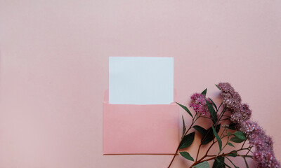 Pink envelope with a white mockup sheet and pink flowers next to it on pink background.