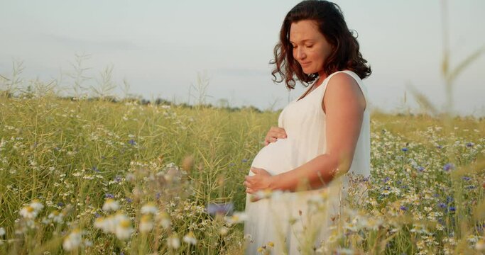 Pregnant woman with a big belly feeling happy outdoors. Adult expecting mother stroking belly on nature.  Middle age white pregnant woman touching her belly while sunset. Woman pregnancy concept.