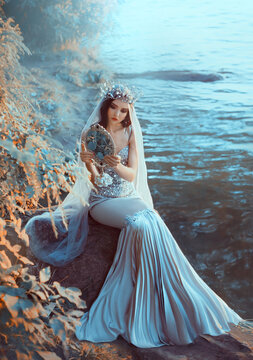 Fantasy woman queen sits on banks of river holding mirror in hands looks at reflection. Silver gray mermaid dress crown on head veil. Autumn nature orange leaves trees blue water. Girl fairy princess