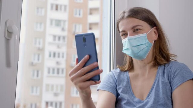 A woman in a protective mask is holding a smartphone and talking on a video call. The patient at the clinic uses her mobile phone to communicate with her friends online.