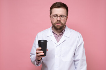 Doctor in a white coat and glasses is very tired and looks sleepy, he holds a paper cup of coffee in hands and stands with eyes closed. 