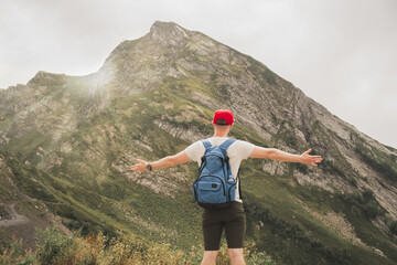 Fototapeta na wymiar A man travels the world and enjoys tourism. The guy spread his arms to the sides and feels freedom in the mountains. A man in a red cap and with a blue backpack meets the dawn