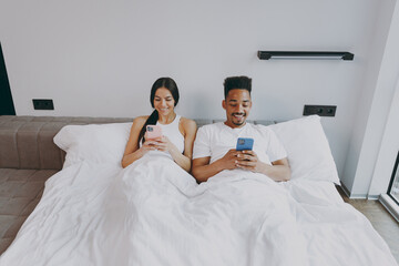 Top view full size body length young couple two family man woman in casual white clothes lying in bed hold use mobile cell phone read chat text spend time together in bedroom lounge home room house
