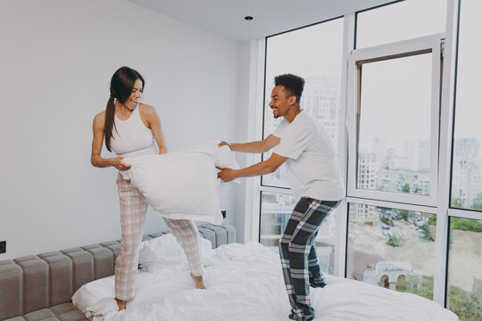 Full Size Body Length Wild Mad Young Couple Two Friends Man Woman Family In Casual White Clothes Stand On Bed Do Pillow Fight Relax Spend Time Together In Bedroom Lounge Home House Wake Up Good Day.