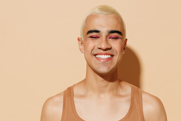 Young smiling fun happy blond latin american hispanic gay man with make up closed eyes in beige tank shirt looking camera isolated on plain light ocher background studio People lgbt lifestyle concept - obrazy, fototapety, plakaty
