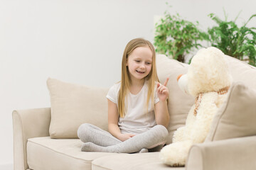 Photo of little girl in white clothes playing on white sofa with teddy bear.