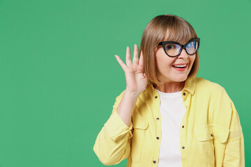 Elderly smiling curious nosy happy caucasian woman 50s in glasses yellow shirt try to hear you overhear listening intently isolated on plain green background studio portrait People lifestyle concept.