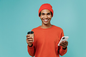 Young happy african american man in orange shirt hat hold takeaway delivery craft paper brown cup coffee to go use mobile cell phone isolated on plain pastel light blue background studio portrait