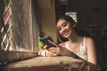 A young smiling asian woman busily chatting on her phone while hanging out in a dimly lit room.