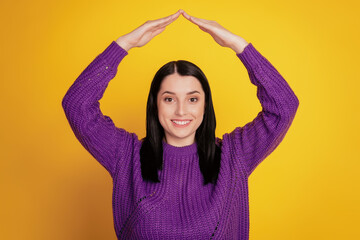 Young smiling happy girl holding folded hands above head like roof home house relocation yellow background studio portrait