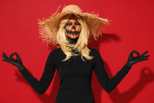Young woman with Halloween makeup mask wears straw hat black scarecrow costume hold hands yoga om aum gesture relax meditate isolated on plain red background studio Celebration holiday party concept.