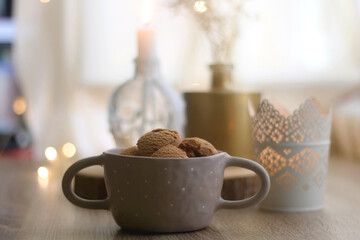 Bowl of cinnamon cookies, lit candles and vase with gypsophila flowers on the table. Selective focus, fairy lights in the background. Hygge at home.