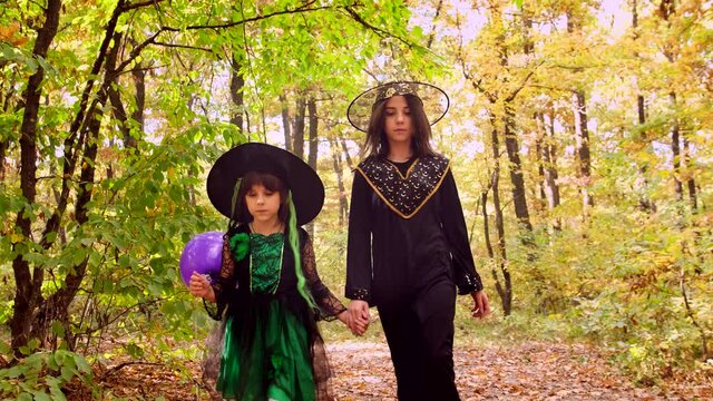 Two girls hold hands in Halloween costumes and hats on their heads holding violet balloon walking into the autumn forest.
